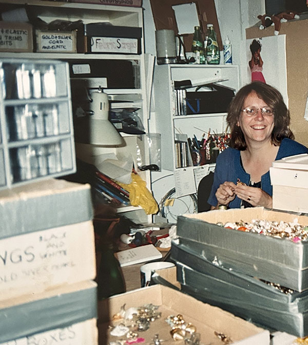 Julie Mollins smiling among stacks of labeled costume jewelry storage boxes in a cluttered backstage workroom.
