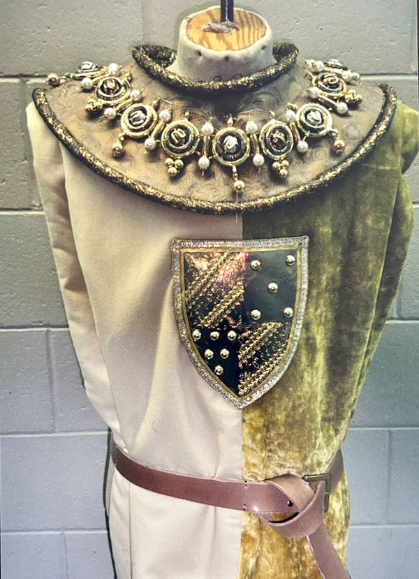 Ornate gold collar with spiral medallions and pearls, worn over a two-toned green and cream tunic with a heraldic shield badge.
