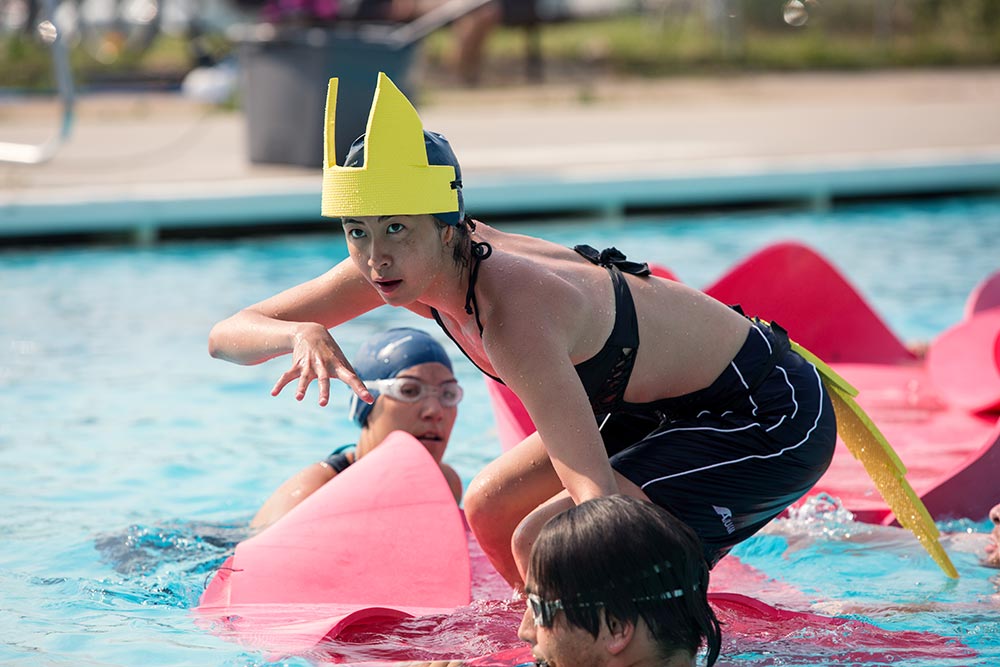 A young person in a black swimsuit and shorts balances on a pink floating mat in a swimming pool, wearing a yellow foam costume resembling a crown or helmet with fins on the back. Two other participants, also in swimwear and goggles, are in the water around them, suggesting a playful or theatrical water activity.